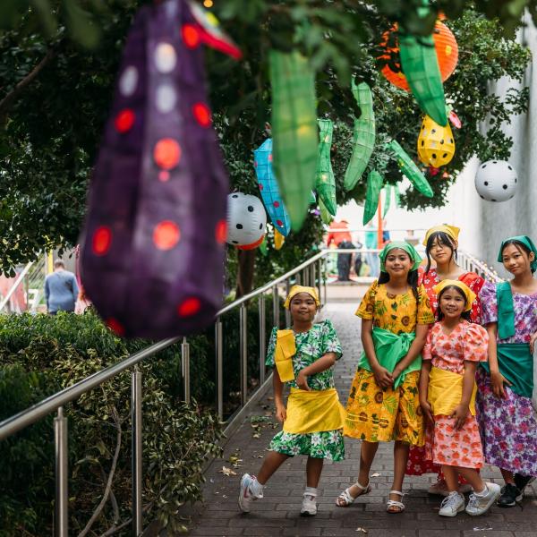Group of children in colourful traditional outfits standing together on a garden path decorated with lanterns.