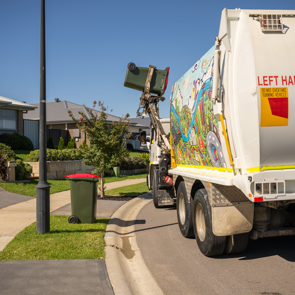 Council garbage truck empties rec bin contents into truck. Another red bin is on the sidewalk. 