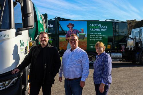 Three people stand in line smiling with large waste trucks in background with FOGO branding.