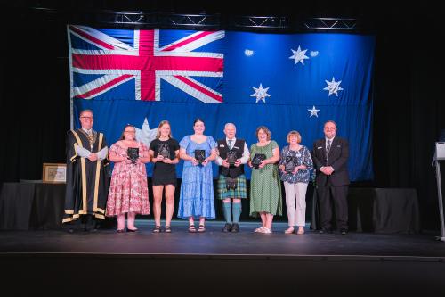 Group of individuals standing on stage with mayor and councillor holding awards in front of an Australian flag.