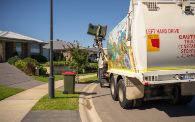 Council garbage truck empties rec bin contents into truck. Another red bin is on the sidewalk. 