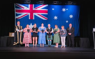 Group of individuals standing on stage with mayor and councillor holding awards in front of an Australian flag.